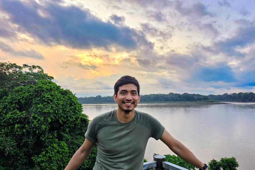 A smiling man stands on a platform above a body of water,  forests, and a stunning sky of clouds in the background.