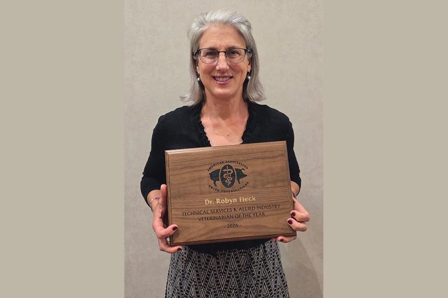 A woman with gray shoulder length hair is wearing glasses, a black v-neck top and gray tweed skirt. She is holding an award plaque.