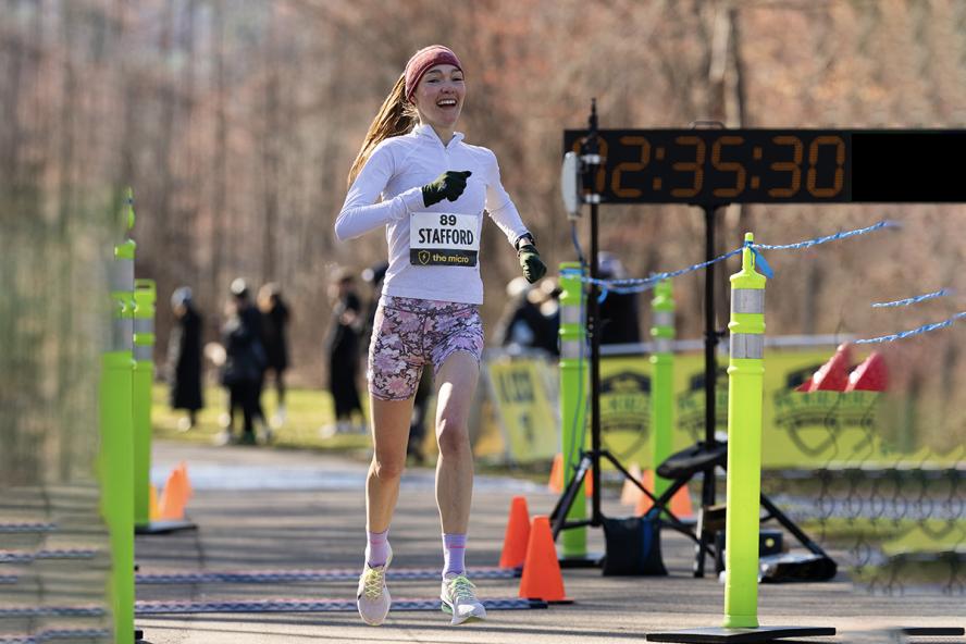 A smiling female runs outside between race cones with a large digital clock in the background that reads “02:35:30.”