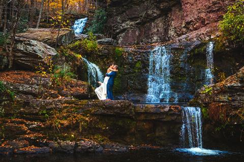 A couple in wedding attire embraces in front of a waterfall. 