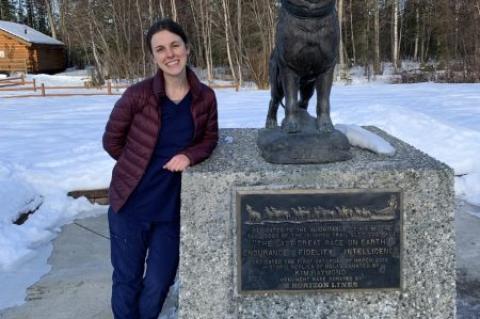 Women standing next to the Iditarod Race monument with a statue of a sled dog on top