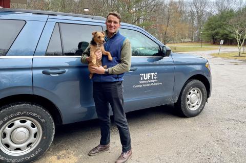 A man wearing a blue vest holding his small dog standing in front of a blue SUV with the Tufts logo on the side door