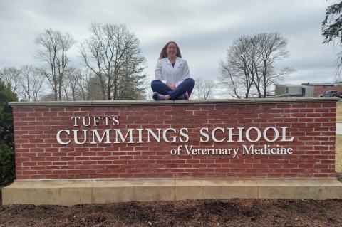 A woman wearing a white coat and blue jeans sits atop a brick wall with sign attached that reads Tufts Cummings School of Veterinary Medicine.