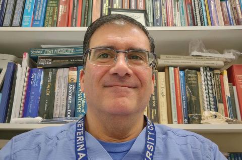 man wearing glasses and blue hospital scrub smiling and stading in front of shelves of books