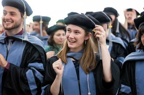 Cummings School doctor of veterinary medicine graduates wearing blue and black gowns and black barets. A smiling man in front is clapping and a smiling woman with long light brown hair is moving her tassel to the other side of her cap.