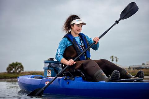 woman with long brown hair pulled up in a pony tail, wearing a white visor and paddling a blue kayak on a lake.