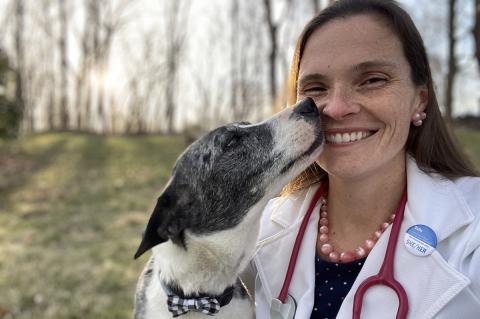 A smiling female veterinarian with long brown hair, wearing a white medical coat and stethoscope around her neck. She is holding a black and white dog that is licking her cheek.