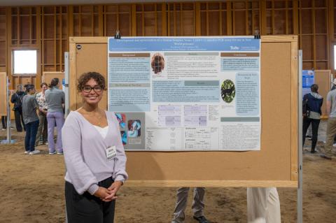 a smiling individual with glasses and curly dark hair wearing a light purple sweater stands beside a research poster.
