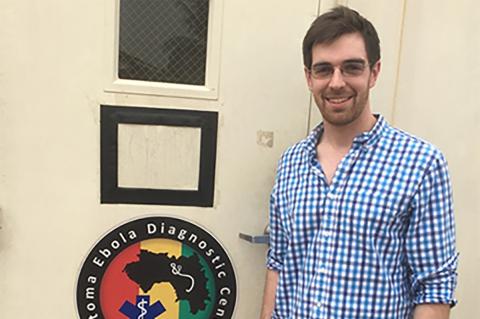 a smiling person wearing glasses, a blue checkered shirt, brown pants and has short brown hair and