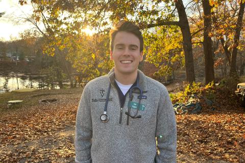 A smiling person with short brown hair wearing a grey 3/4 zip pullover and stethoscope draped around their neck. They are standing outside surrounded by fall foliage.