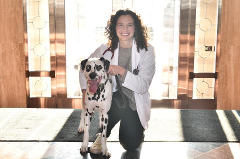 a smiling individual with dark hair wearing a white lab coat and a stethoscope around their neck kneels next to a Dalmatian