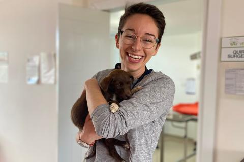 A smiling student with short brown hair dressed in a long sleeve gray shirt wearing glasses and holding a small brown puppy with white paws