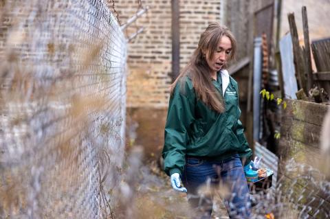 An individual with long brown hair wearing a green jacket, blue jeans, and rubber gloves carries a container of supplies in one hand while walking beside a chain-link fence