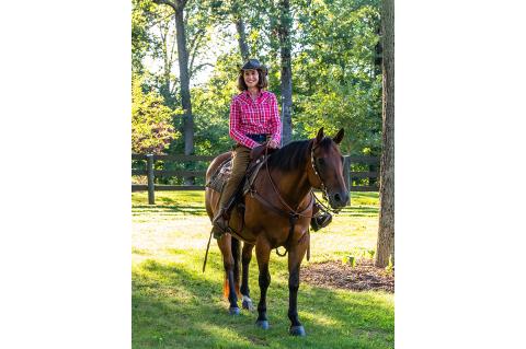 A smiling individual wearing a pink and red checkered cowboy shirt and a cowboy hat sitting on a horse outdoors on a sunny day