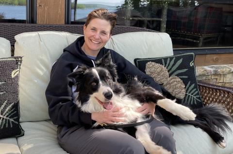 A person smiling wearing gray pants and a black sweatshirt sits on an outdoor couch with a black and white dog