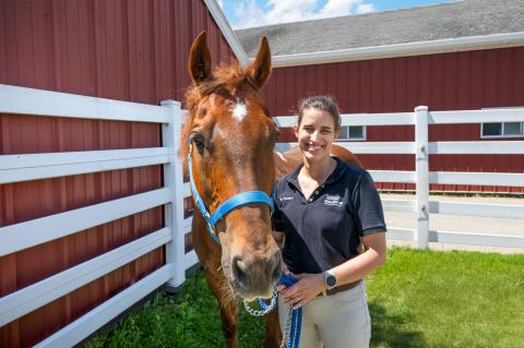Photo of a person posing with a brown horse outside in a holding pen with a red barn in the background.