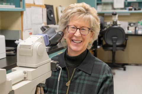  Individual with glasses smiling and sitting at a microscope.