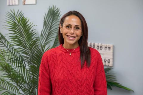 Person smiling in a red sweater with long dark hair standing in front of a green plant.