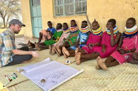 People in Kenya sitting on a mat outside against a yellow building while another person provides educational instruction.