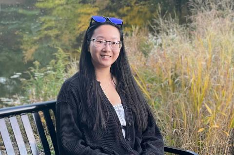 Individual smiling with long dark hair and glasses sitting on a bench outdoors.