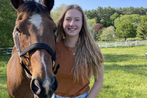 Person standing outside with long blonde hair next to a brown horse on a sunny day.
