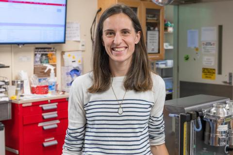 A person with medium-length brown hair, smiling, is standing in a laboratory, wearing a striped sweater.