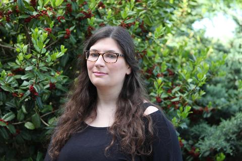 an individual with long dark hair wearing glasses and a black shirt standing outdoors in front of a green bush with red berries.