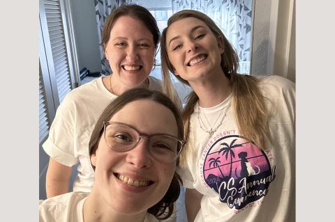 Three individuals standing close together smiling wearing white and pink short sleeve t-shirts.
