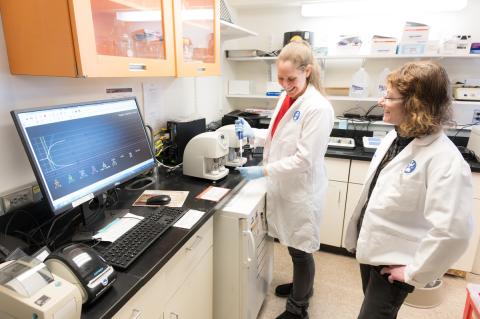 Two smiling individuals interacting in a lab and both wearing white lab coats.