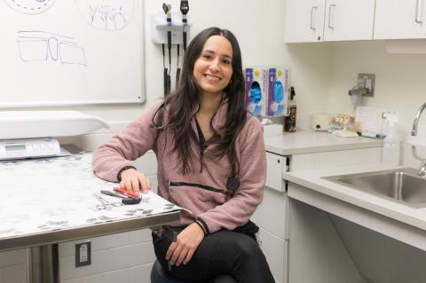 A person with long dark hair sits smiling in an exam room with black pants and a mauve top on.