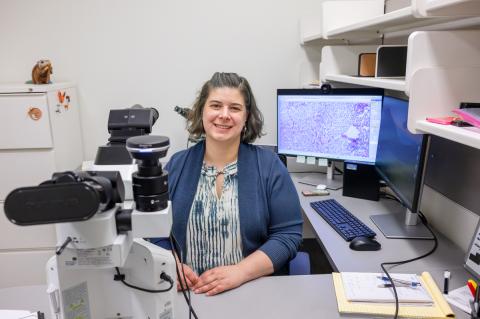 Dr. Laura White (she/her) sitting in her office in front of a large microscope.