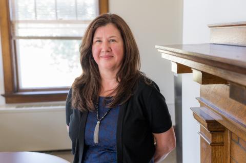 Portrait of person with long brown hair, blue dress, black jacket, and necklace standing in an office.