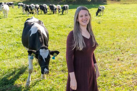 Megan Rokop, Assistant Dean for Community Engagement and Outreach, standing and smiling in front of a grassy pasture with several black and white dairy cows grazing in the background on a sunny day.