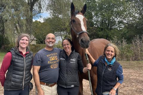 Four individuals standing outside next to a brown and white horse at a stable.