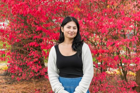 Cara Kaminski, a D.V.M./Master of Public Health student, stands outdoors in front of a bush with vibrant red leaves, wearing a black top, light cardigan, and denim skirt, smiling at the camera.