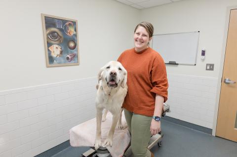 A smiling person stands beside a large light-colored dog on an exam table in a veterinary clinic room. The dog faces the camera with its mouth slightly open. The room has white tiled walls, a medical poster showing an eye diagram, a whiteboard, and a door in the background.