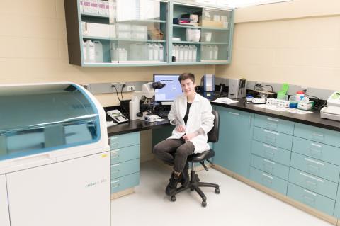 a person wearing a white lab coat sits on a stool in a laboratory, smiling at the camera, with a microscope, computer monitor, and research equipment on surrounding countertops.