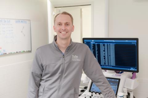 A person in a gray Cummings School of Veterinary Medicine at Tufts University jacket stands beside a computer workstation in a clinical setting, with a whiteboard in the background.