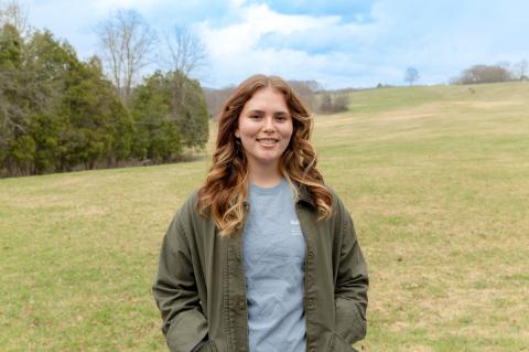A young woman smiling while standing in a grassy field with trees and blue sky behind her.