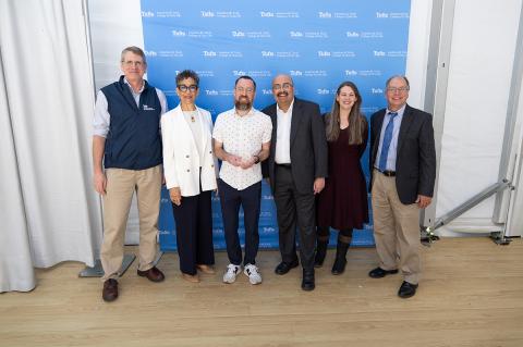Graduate poses with award alongside five deans and president in front of blue Tufts College of Civic Life sign.
