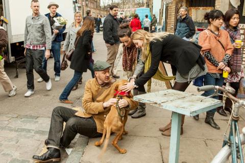 A man sitting a sidewalk curb with his dog, as two young women pet the dog, and other people look on. Research shows that walking dogs isn’t just exercise, it also helps create social bonds in communities