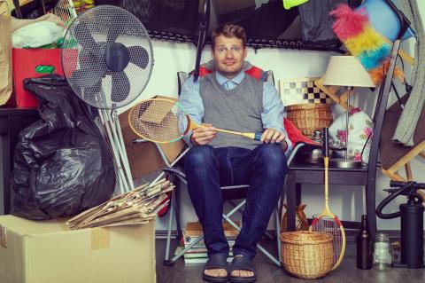 A man sits in a garage surrounded by clutter.